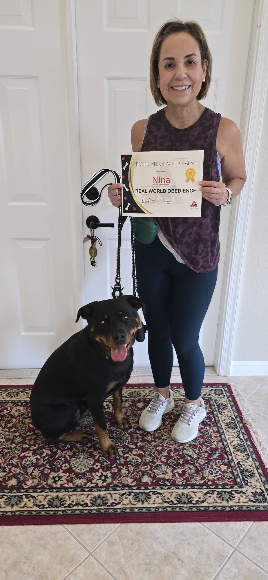 A client holding Nina's Real World Obedience certificate with her Rottweiler sitting calmly on leash.