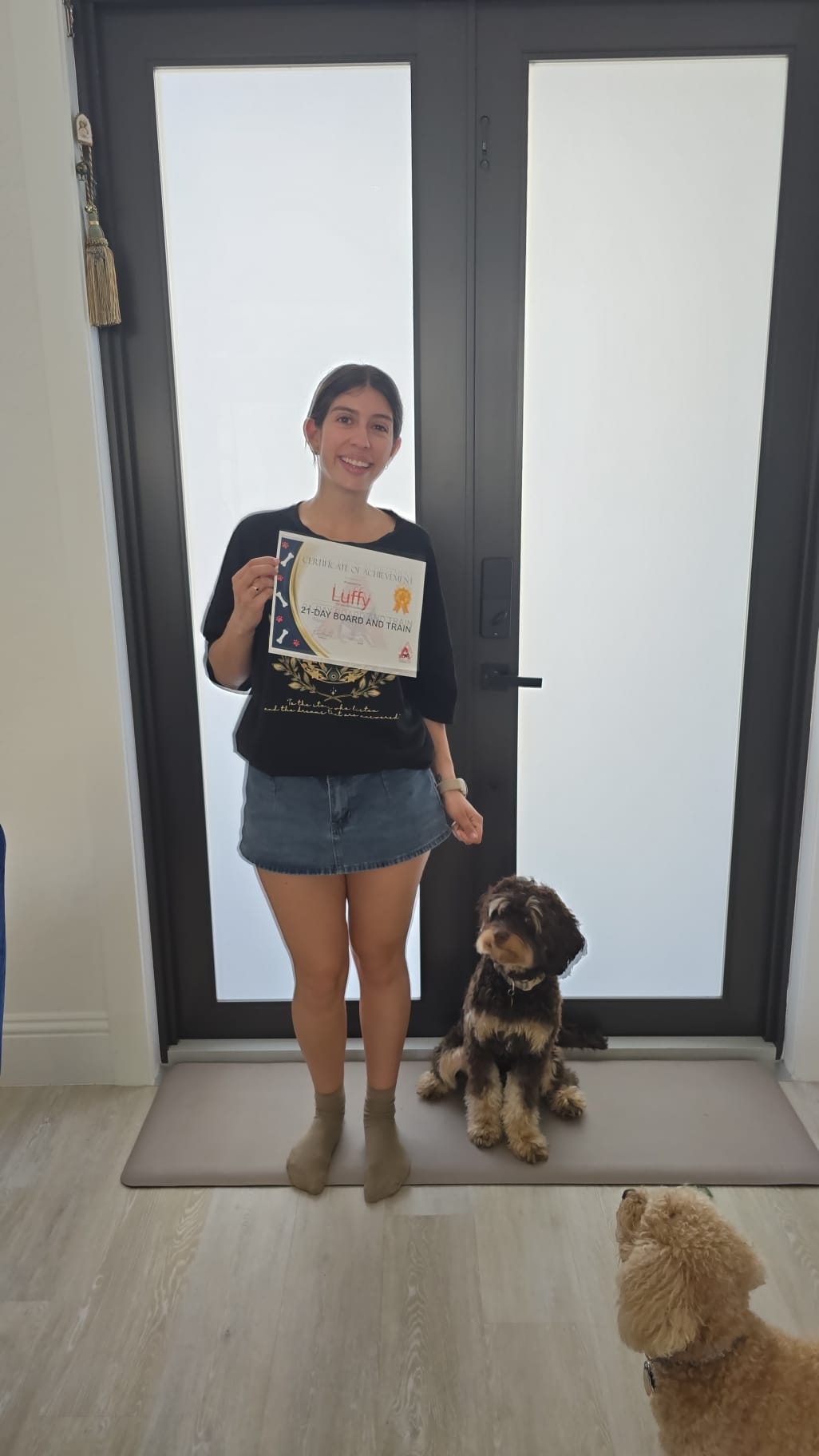 A client standing by the front door holding Luffy's 21-Day Board and Train certificate with her dog sitting beside her.