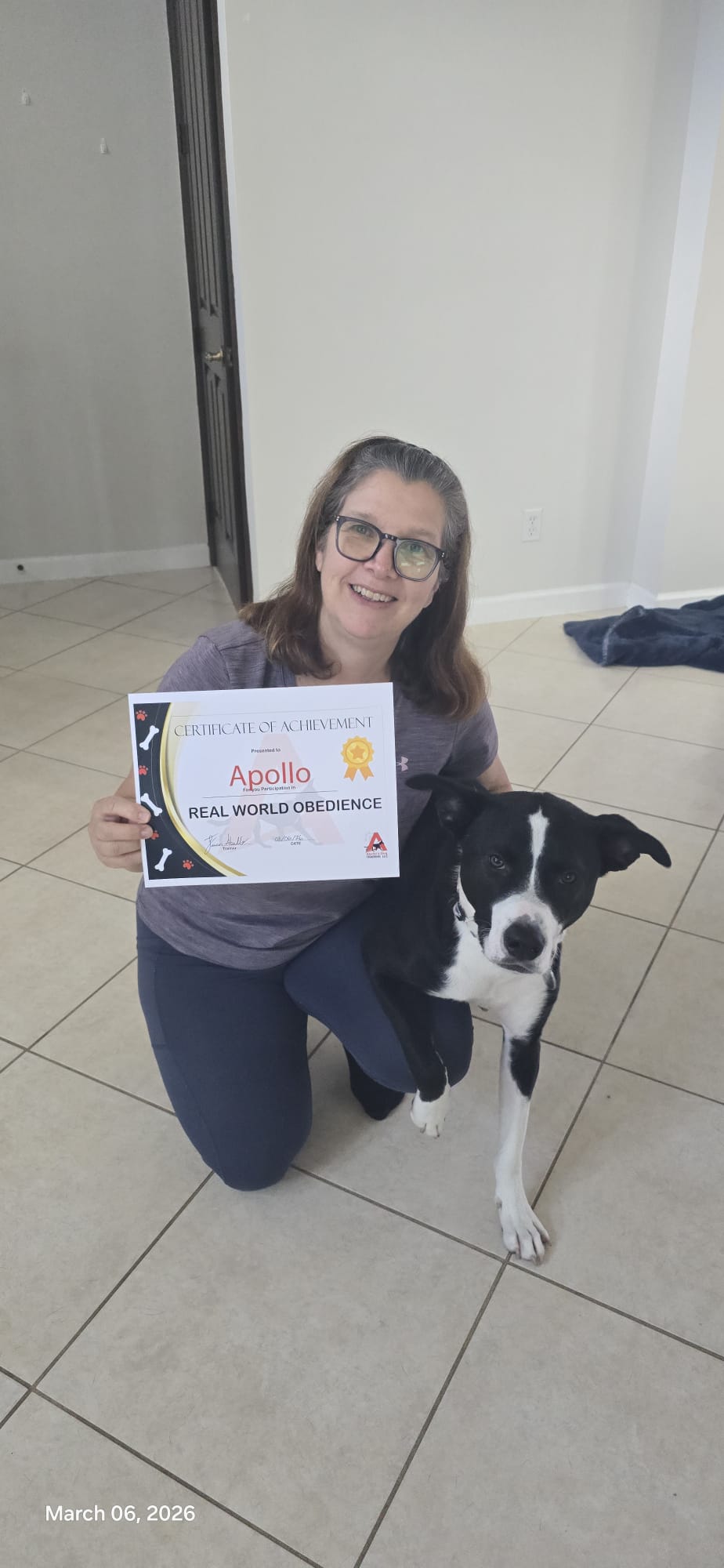 A client kneeling beside her black and white dog while holding Apollo's Real World Obedience certificate.