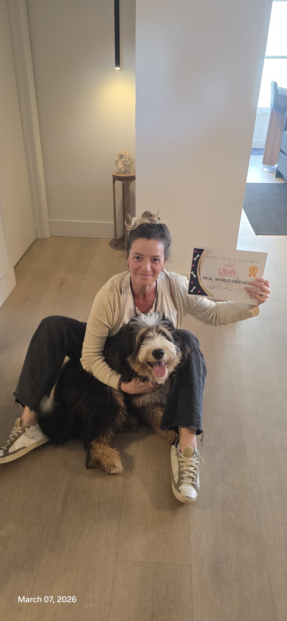 A client sitting on the floor hugging her Bernedoodle while holding his certificate.